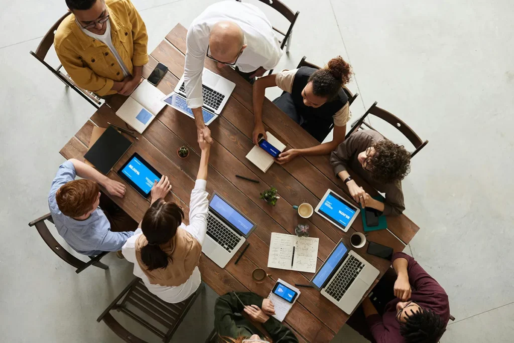 People meeting at a table with iPads, laptops, and notebooks. Two people shaking hands.